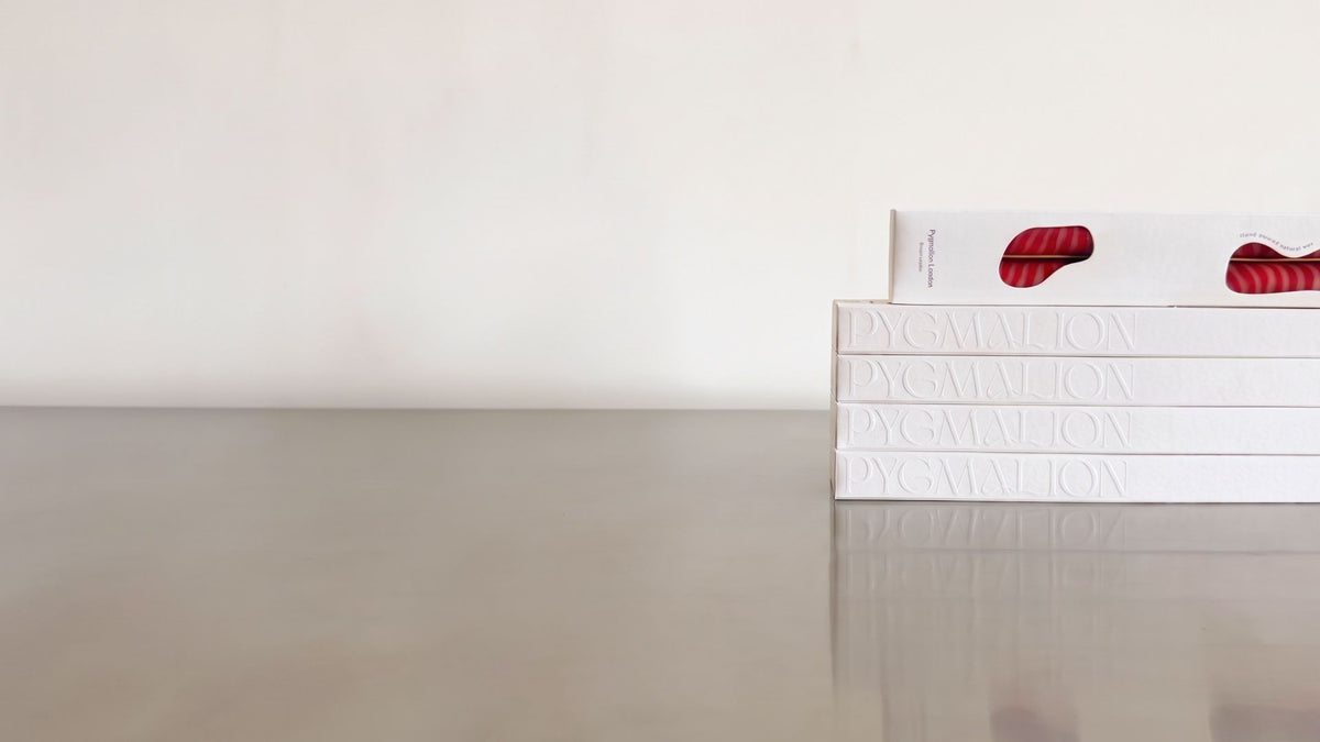 A stack of white Pygmalion gift boxes with the name embossed on each side, topped by a box whose die-cut windows show red and white striped dinner candles.